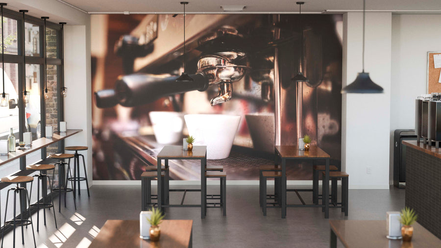 A modern cafe interior with wooden tables, stools, and potted plants features natural light from large windows and pendant lights. The back wall displays the Espresso Machine Wall Mural showing a close-up of an espresso machine with white cups.