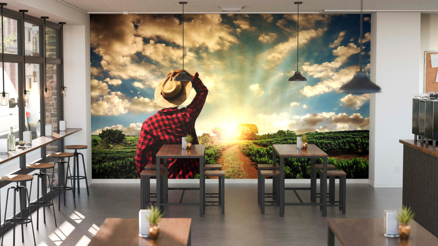 A cozy modern café with wooden tables and stools. The back wall showcases the "Farmer's Morning View Wall Mural," depicting a person in a plaid shirt and hat watching a sunrise over fields, while small plants decorate the tables.