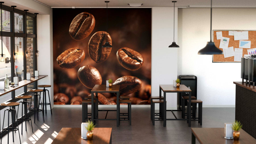 A modern cafe interior with wooden tables, stools, potted plants, large windows, pendant lights, and a bulletin board. The Floating Beans Wall Mural (peel and stick) covers one wall with images of floating coffee beans.