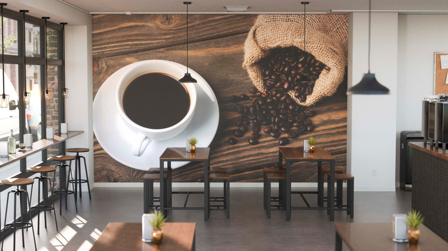 A modern coffee shop interior with wooden tables, black chairs, and potted plants features the Fresh Brew and Beans Wall Mural on the back wall. Pendant lights hang above as sunlight streams in through tall windows.