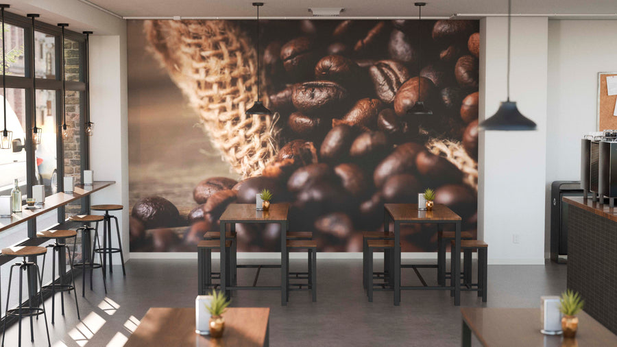 A modern café interior with wooden tables and stools, small plants, pendant lights, and a large Fresh Coffee Roast Wall Mural featuring coffee beans. Floor-to-ceiling windows on the left bring in natural light.