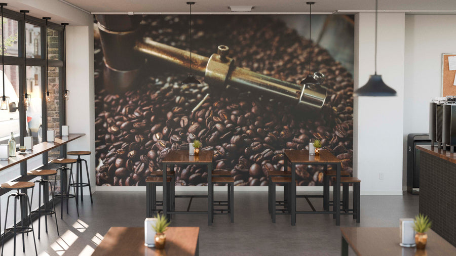 A modern café featuring wooden tables, black stools, and potted plants on each table. The back wall displays the Fresh Roast Wall Mural showing coffee beans and roasting gear, with pendant lights above and windows to the left.
