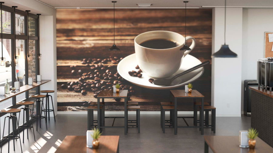 A modern cafe interior with wooden tables, black chairs, potted plants, and large windows features the Freshly Brewed Black Coffee Wall Mural—a close-up of a white cup of coffee with beans on rustic wood—plus pendant lights overhead.
