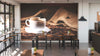 Modern coffee shop interior with wooden tables, black chairs, and potted plants. The "Freshly Brewed Delight Wall Mural" displays a white coffee cup on coffee beans with burlap background. Pendant lights hang from the ceiling.