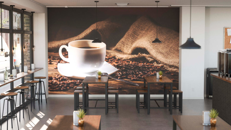 Modern coffee shop interior with wooden tables, black chairs, and potted plants. The "Freshly Brewed Delight Wall Mural" displays a white coffee cup on coffee beans with burlap background. Pendant lights hang from the ceiling.