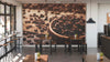 Modern cafe interior with wooden tables and stools, small plants on each table, and a Freshly Roasted Wall Mural showcasing coffee beans in a wooden scoop. Pendant lights hang from the ceiling, with large windows along the left wall.