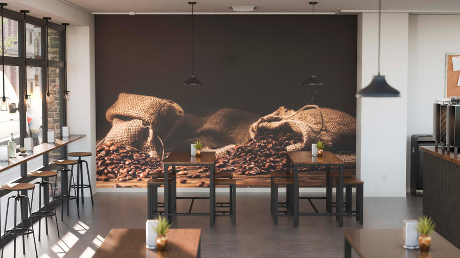 A modern coffee shop features wooden tables and stools, potted plants on each table, pendant lights above, large windows for natural light, and a Freshly Roasted Beans Wall Mural of burlap sacks spilling roasted coffee beans.