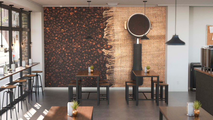 A modern coffee shop with wooden tables, stools, potted plants, and the "Grounded in Flavor Wall Mural" featuring coffee beans and a portafilter on burlap. Pendant lights hang above; natural light beams through tall windows on the left.
