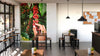 A bright coffee shop features wooden tables, stools, pendant lights, large windows, potted plants, a corkboard with notes, and a modern vibe. The Handpicking Coffee Cherries Wall Mural adds an inviting touch with its custom design of hands picking red cherries.