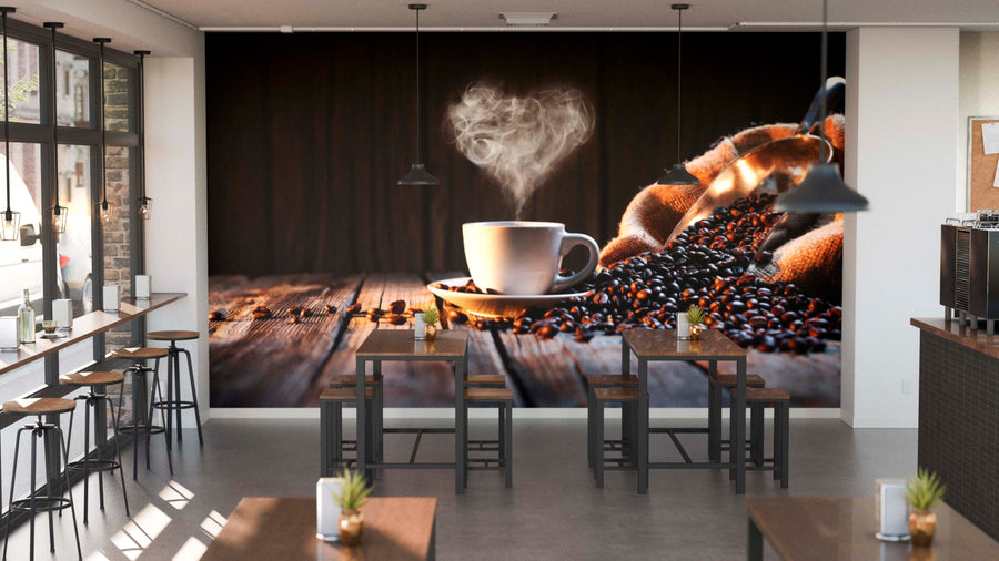 A modern café interior with wooden tables, industrial pendant lights, potted plants, and a cozy vibe. The back wall is adorned with the Heartwarming Coffee Wall Mural showing a steaming coffee cup on beans and a striped cloth on rustic wood.