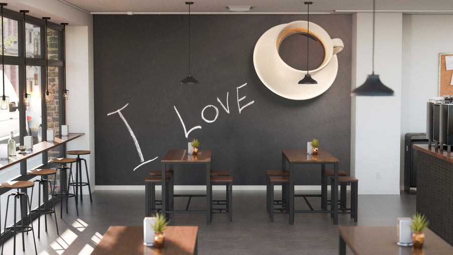 A modern café with wooden tables and stools features the I Love Coffee Wall Mural on a black wall, showing a large coffee cup and “I LOVE.” Small plants decorate each table, while pendant lights add to the cozy atmosphere.