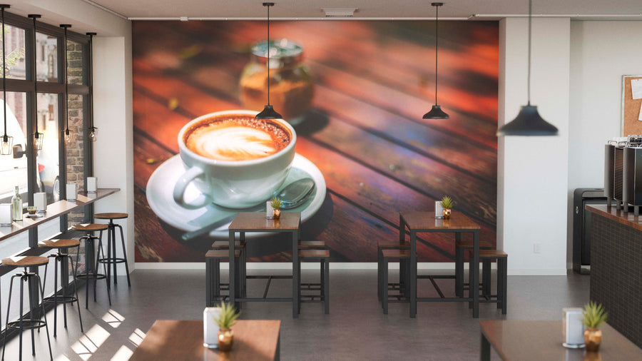 A modern café with wooden tables, black stools, and hanging lights showcases the Morning Coffee Wall Mural—a large image of a cappuccino in a white cup on wood. Small plants complete the inviting, charming atmosphere.