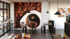 A modern café with wooden tables, stools, potted plants, and large windows features the Natural Coffee Wall Mural depicting a coffee cup and beans on the back wall. Pendant lights hang above and a corkboard decorates the right wall.