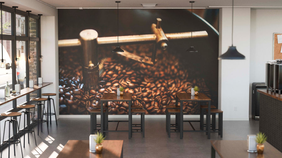 A modern café with wooden tables, black stools, and potted plants on each table features large windows and the Perfect Batch Wall Mural of coffee beans and roasting gear on the back wall, highlighting the coffee theme.