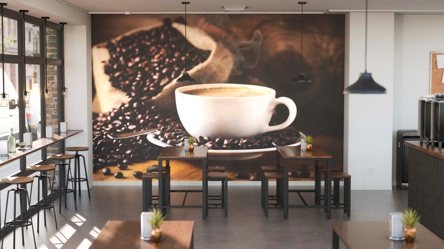 A modern café with wooden tables and stools features the Perfect Cup Wall Mural of a steaming coffee cup and beans. Potted plants, pendant lights, and tall windows brighten the sunlit space with natural light.
