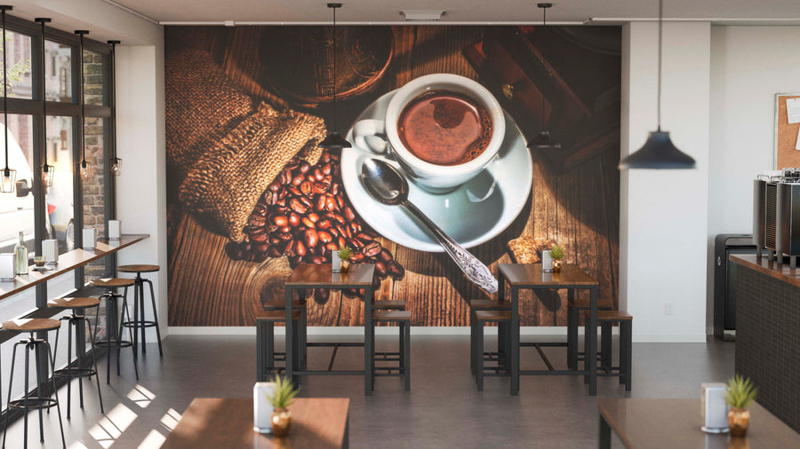Modern café interior with wooden tables, stools, potted plants, and large wall art featuring the Picking Coffee Wall Mural peel and stick design. Pendant lights hang overhead for a warm ambiance.