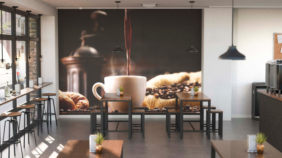 A modern coffee shop with wooden tables, stools, potted plants, and large windows features the Pouring Fresh Coffee Wall Mural on the back wall, depicting a coffee cup being filled with beans and a grinder for a warm, inviting vibe.