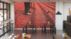 A modern cafe with large windows and wooden tables features the vibrant "Raking Ripe Coffee Cherries Wall Mural" showcasing legs, shoes, and drying red coffee cherries. Small plants and hanging lights add warmth to the inviting space.
