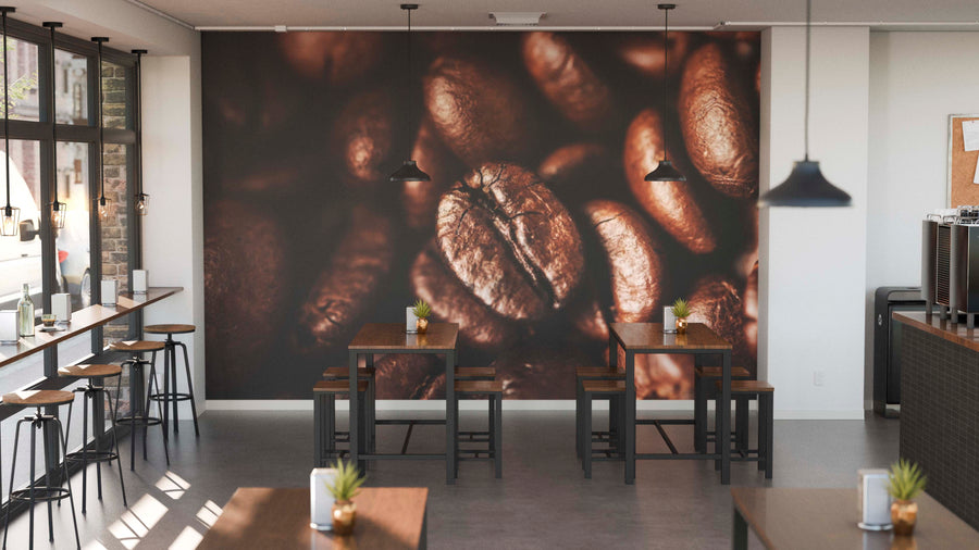A modern cafe with wooden tables and stools is empty. The back wall features the Roast Detail Wall Mural of coffee beans. Sunlight streams through tall windows, highlighting potted plants on tables and black pendant lights overhead.