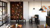 A modern café with wooden tables and stools features the Roasted Coffee Beans Wall Mural, hanging pendant lights, a bulletin board with papers, small potted plants on tables, and large windows that fill the space with natural light.