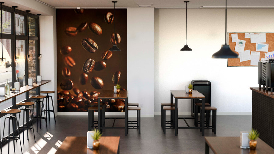 A modern café with wooden tables and stools features the Roasted Coffee Beans Wall Mural, hanging pendant lights, a bulletin board with papers, small potted plants on tables, and large windows that fill the space with natural light.