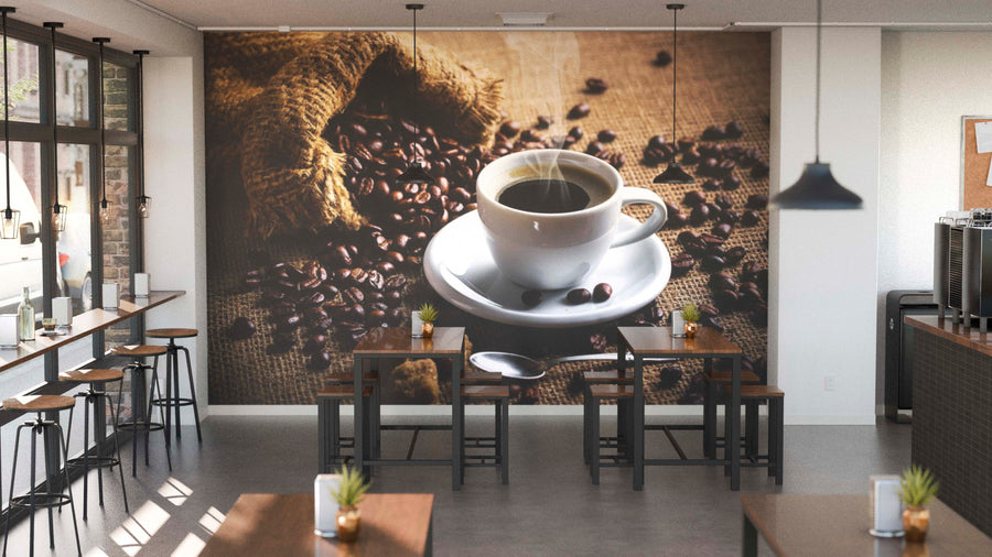A modern café interior with wooden tables, potted plants, and large windows. The back wall displays the Steaming Cup of Coffee Wall Mural featuring a steaming coffee cup and beans spilling from a burlap sack.