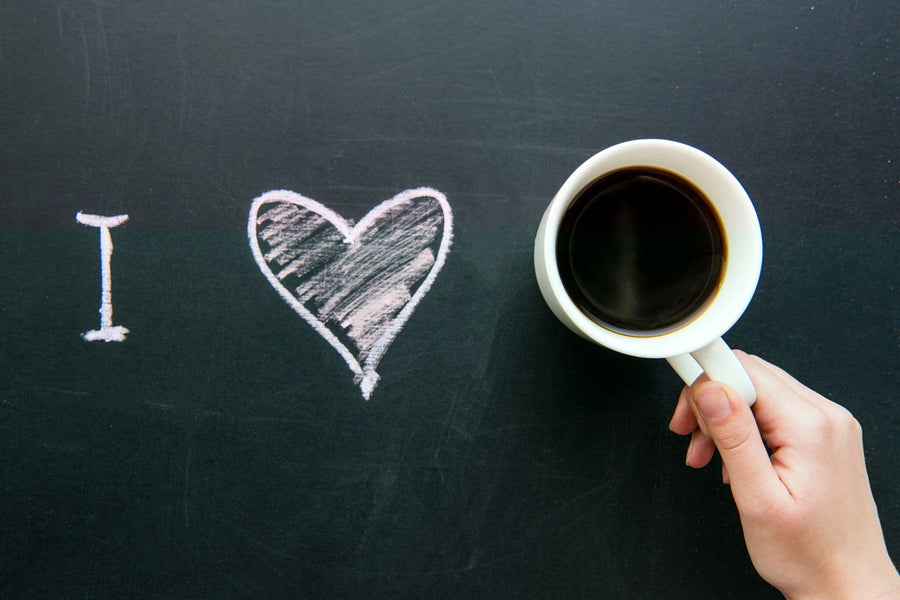 A modern café with wooden tables and potted plants features the Coffee is Love Wall Mural—a large chalk-style “I ♥️” and hand with a coffee cup—illuminated by daylight from big windows in the empty space.