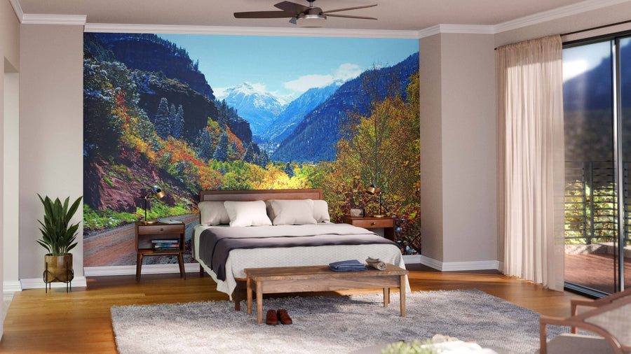 A modern bedroom showcases the Colorado Mountains Autumn Road Wall Mural behind the bed, with a beige color palette, wooden furniture, gray rug, plant, and floor-to-ceiling windows draped in sheer curtains.