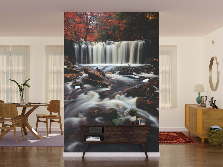 A living room is enhanced by the Colorful Foliage Wall Mural, showcasing an autumn forest waterfall. Flowing water, red and green leaves, modern furniture, a dining table, lush plants, and a round mirror complete the inviting space.