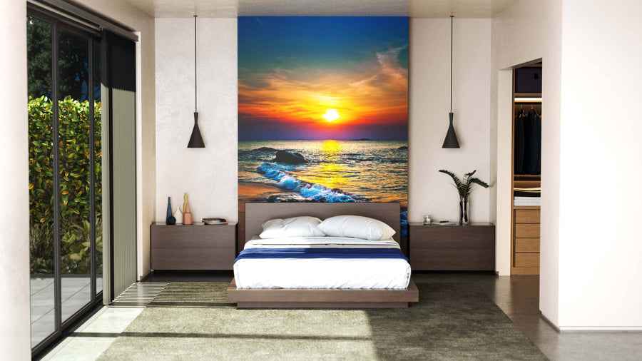A living room with pale green walls features the Colorful Sunset Over the Sea Wall Mural—a vibrant peel-and-stick beach sunset with waves, rocks, and a colorful sky. Wooden furniture, plants, a red rug, and gentle natural light complete the space.