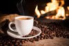 A steaming white coffee cup rests on a saucer amid scattered beans on a rustic wooden table adorned with  Cozy Coffee Wall Mural. The blurred flames of a cozy fireplace create a warm ambiance, while a burlap sack is partially visible on the left.