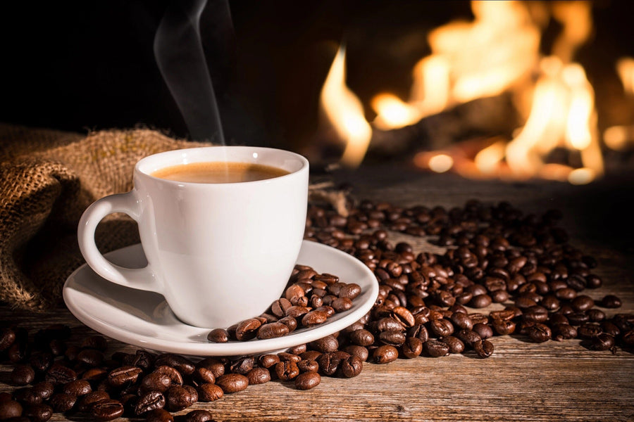 A modern café with wooden tables, stools, potted plants, and large windows. The back wall features the Cozy Coffee Wall Mural—a steaming coffee cup on beans by a warm fire—adding a cozy vibe to the space.