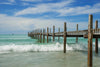 The Crashing Waves Wall Mural transforms a serene coastal scene into art, showcasing a long wooden pier in the turquoise sea under partly cloudy skies, with gentle waves creating white foam and a distant boat adding depth to this stunning vista.