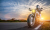 A motorcyclist wearing a helmet rides a cruiser motorcycle on an open road under the sun's warm glow and dramatic cloud patterns, resembling the "Cruising into Sunset Wall Mural," with the headlight on as they lean right, chasing the endless horizon.