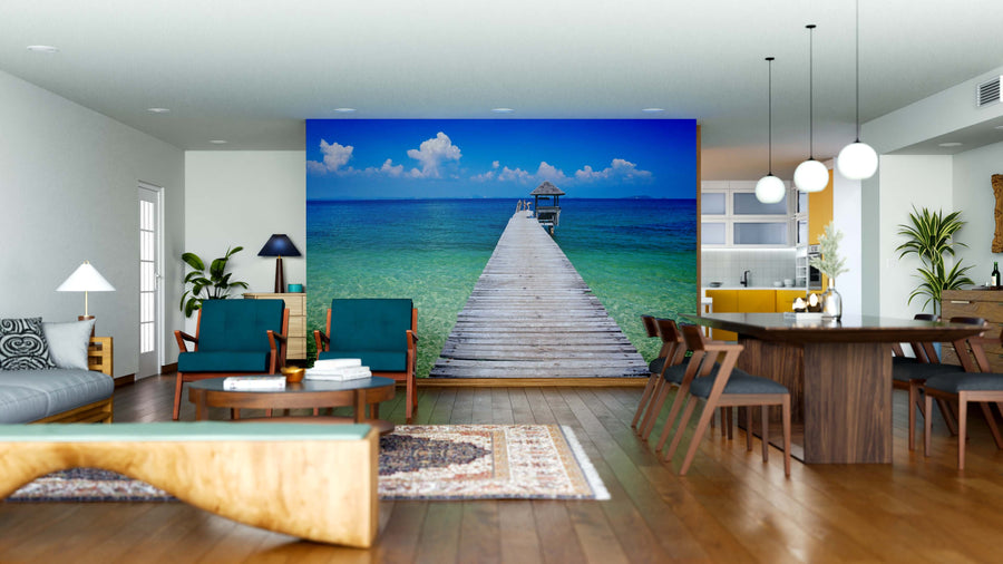 A modern open-plan living and dining area features wooden furniture, teal chairs, and the Crystal Blue Pier Wall Mural—a peel and stick mural showing a wooden pier stretching into turquoise ocean waters under a bright blue sky.
