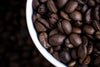 Close-up of glossy dark brown coffee beans in a white bowl, set in the upper right corner with blurred beans behind. Ideal for decorating your space with the Cup of Coffee Beans Wall Mural—available as custom prints or peel and stick murals.
