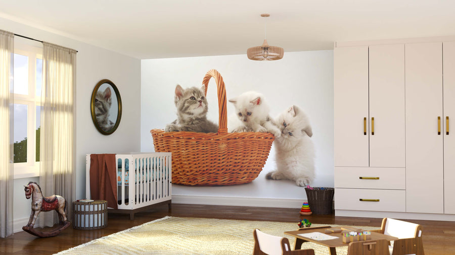 A cozy nursery features a crib, rocking horse, and toys. One wall displays the Curious Little Cats Wall Mural—a playful peel and stick design showing three kittens in a basket. Sunlight streams through sheer curtains, with a circular mirror reflecting the space.