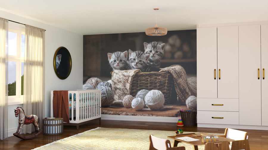 A cozy nursery with a crib, rocking horse, and toys features the Curious Tabby Trio Wall Mural showing three gray tabby kittens in a basket with yarn. Sunlight streams onto light wood floors, a soft rug, and white furniture with gold handles.