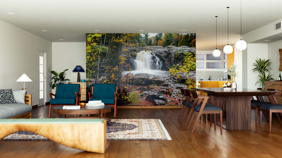 A modern open living and dining area with wood flooring, blue armchairs, and a dining table features the Dead River Falls Wall Mural—a peel and stick forest waterfall scene. Kitchen, houseplants, and pendant lights complete the vibrant space.