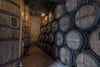 In a dimly lit cellar, with stone walls adding rustic charm, rows of stacked wooden barrels line the right, and a large wooden tank sits on the left. A custom Wine 19 Wall Mural enhances the space as shadows from a single bulb amplify the ambiance.