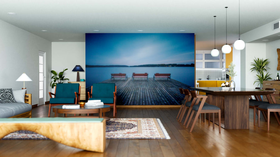 A modern open-plan living and dining area features teal chairs, a beige sofa, a wooden coffee table, and plants. The Dock with Benches Wall Mural—a peel and stick design—adds tranquil lake views, enhanced by pendant lights.
