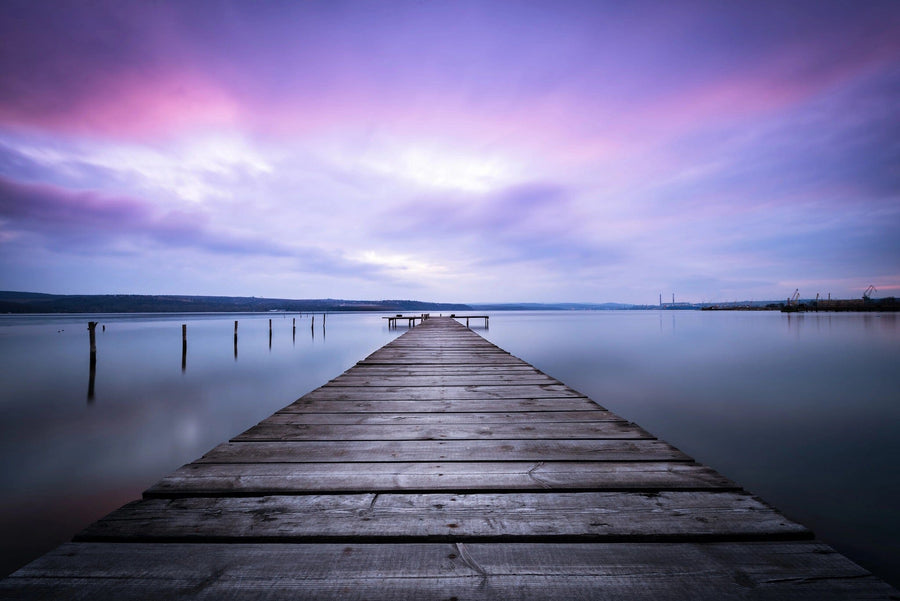 A modern open-plan living and dining room with wooden furniture, green chairs, hanging lights, plants, and the Dreamscape Pier Wall Mural depicting a serene dock under a purple-blue sunset sky.