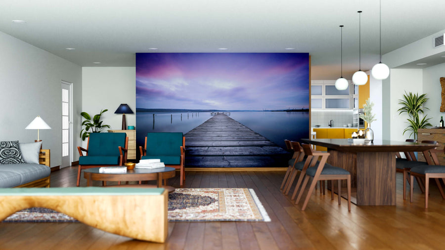 A modern open-plan living and dining room with wooden furniture, green chairs, hanging lights, plants, and the Dreamscape Pier Wall Mural depicting a serene dock under a purple-blue sunset sky.