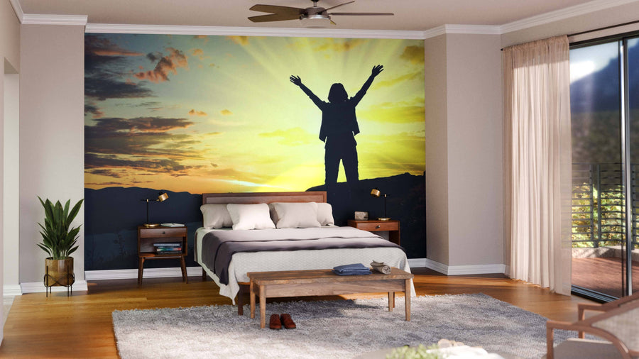 A modern living and dining area with wood flooring features the Dusk Wall Mural, showing a person with raised arms at sunset. Contemporary furniture, pendant lights, and houseplants add comfort with natural tones and textures.