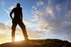 A silhouetted figure on rocky terrain under a vast sky painted with blue, orange, and yellow hues is beautifully captured in the Embracing the Horizon Wall Mural, showcasing nature's artwork in vivid detail.