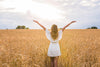 A woman in a white dress with arms outstretched stands in a golden wheat field under a luminous sky, embodying the serenity of the Inspirational 4 Wall Mural. Her back is to the camera, facing bright clouds and distant trees as the sun peeks through.