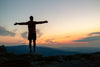 A person stands on a rocky hill, arms open to a vibrant sunset, reminiscent of the Inspirational 6 Wall Mural. The sky shines in orange, pink, and blue hues against distant mountains. They're dressed in shorts and a long-sleeved shirt.