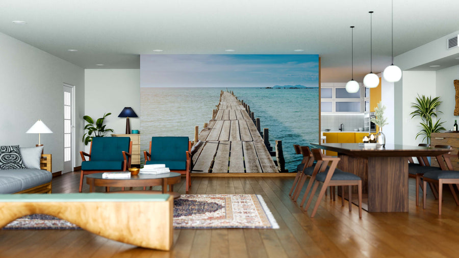A modern open-plan living and dining room features wooden furniture, blue chairs, pendant lights, and the Endless Blue Wall Mural depicting a wooden pier leading into a tranquil blue sea under a clear sky for a serene coastal vibe.
