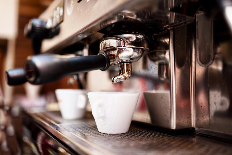A modern cafe interior with wooden tables, stools, and potted plants features natural light from large windows and pendant lights. The back wall displays the Espresso Machine Wall Mural showing a close-up of an espresso machine with white cups.
