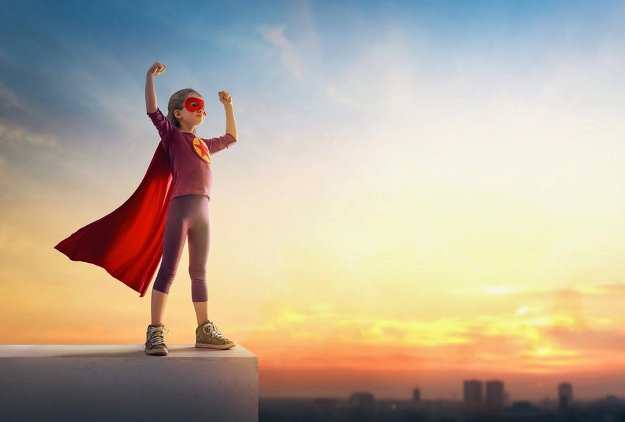 A modern open-plan living/dining room with wood floors, teal armchairs, and the "Fearless and Free Wall Mural" featuring a child in a red cape posing as a superhero against a sunset sky for playful, contemporary style.